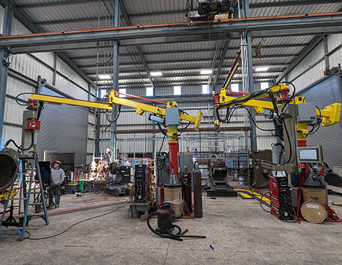 A spacious industrial workshop with high ceilings, featuring several large, yellow robotic arms. Equipment and tools are scattered around, and a person works at one of the stations.