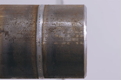 Close-up of the end of a metal pipe with a visible weld seam running horizontally. The pipe has a slightly weathered and rusty appearance, set against a plain, light gray background.